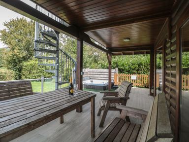 A covered wooden patio with a wooden table and chairs a bottle with two glasses on the table a metal spiral staircase outside and a fenced hot tub in a garden at Ash in Swanage