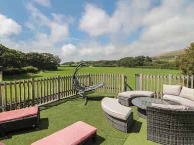An outdoor patio with wicker seating and lounge chairs on artificial grass overlooking green fields and trees at Ash in Swanage