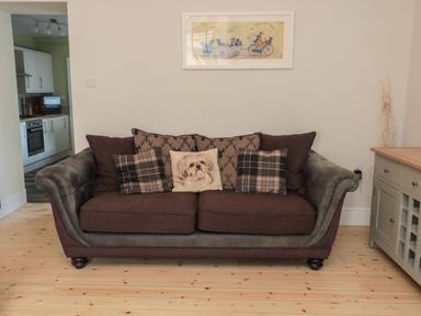 A brown couch with patterned and dog face cushions in a living room with wooden floor and kitchen visible through doorway at 55 Wellwood Street in Amble