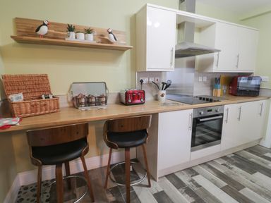 A kitchen with a wooden counter, two wooden chairs, a red toaster, a stove, an oven, white cabinets, and a wooden shelf with two bird figurines and plants at 55 Wellwood Street in Amble