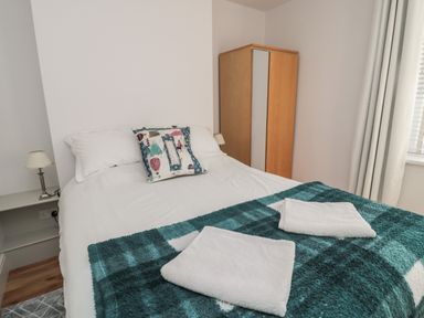 A bedroom with a double bed with white linens and a teal checkered blanket and two folded white towels on the bed beside a wooden wardrobe and a lamp on the bedside table at 55 Wellwood Street in Amble