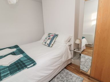 A bedroom with a bed white linens and a patterned pillow beside a wooden wardrobe with a mirror and a small lamp on a wall shelf at 55 Wellwood Street in Amble