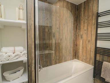 A bathroom with a white bathtub with a glass shower screen wooden panel walls white shelves with towels and a black towel radiator at 55 Wellwood Street in Amble