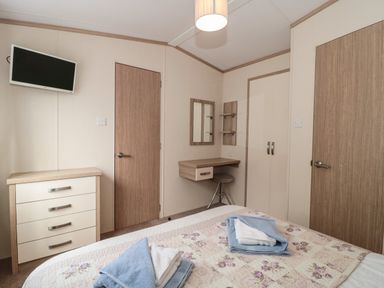 A bedroom with a bed, folded towels on the bed, a chest of drawers, a wall-mounted television, a dressing table with a mirror and stool, and two wooden doors at Beech in Swanage