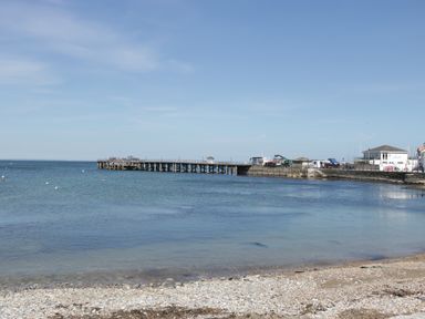 A pebble beach with a wooden pier extending into calm sea water and buildings near the shore at Beech Swanage