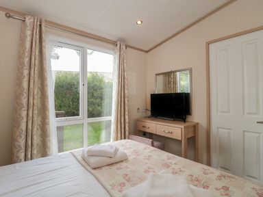 A bedroom with a bed covered with floral blankets and folded towels by a window and a wooden dresser with a TV and mirror at Birch in Swanage