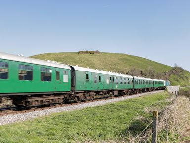 A green passenger train traveling on railway tracks through grassy countryside near a hill at Birch in Swanage