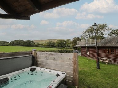 An outdoor hot tub on a wooden deck with a grassy area a wooden cabin a lamppost and hills in the background at Oaks Swanage