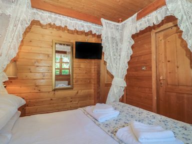 A bedroom with wood panel walls a bed with white sheets folded towels a wall mirror a mounted television and lace curtains at Cherry in Swanage