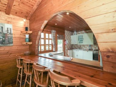 A wooden kitchen bar with four stools and an arched opening to a kitchen with cabinets and a tiled wall at Walnut in Swanage