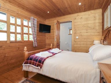 A bedroom with a wooden bed and white bedding a red plaid blanket and towels on the bed a wooden door open to an en suite bathroom and a wall-mounted television at Walnut in Swanage