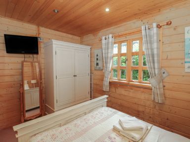 A bedroom with wooden walls and ceiling a bed with folded towels a white wardrobe a standing mirror a window with plaid curtains and a mounted television at Pine in Swanage