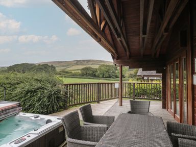 A wooden covered patio with a hot tub and wicker chairs around a table overlooking countryside at Pine in Swanage