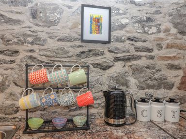 A kitchen counter with a mug rack holding colorful mugs a black electric kettle and three white canisters on a stone wall at The Artist's Barn in Ayle near Cappawhite