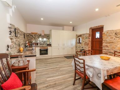 A kitchen with white cabinets wooden chairs a dining table with a patterned tablecloth and stone walls at The Artist's Barn in Ayle near Cappawhite