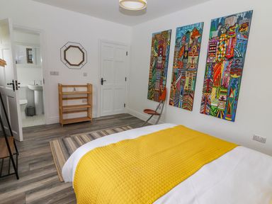 A bedroom with a bed covered by a yellow blanket three colorful paintings on the wall a chair a mirror wooden shelves and an open door leading to a bathroom at The Artist's Barn in Ayle near Cappawhite