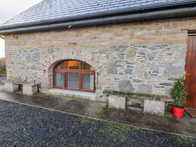 A stone building exterior with two stone benches and a red potted plant near a wooden door at The Artist's Barn in Ayle near Cappawhite