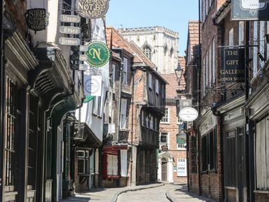 A narrow street with historic buildings and hanging shop signs leading to a large stone church tower in the background