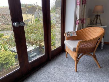 A wicker chair with a cushion and a book on it next to glass doors and a lamp on a side table at Foxglove Barn in South Brent