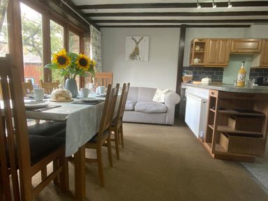 A dining area with a table set with cups and a vase of sunflowers next to a striped sofa and kitchen cabinets at Foxglove Barn in South Brent