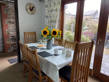 A dining area with a wooden table and six chairs set with a vase of sunflowers and tea set near large wooden framed glass doors at Foxglove Barn in South Brent