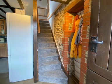 A narrow staircase with carpet leading upstairs next to a white refrigerator and coats hanging on hooks in a brick and stone wall at Foxglove Barn in South Brent