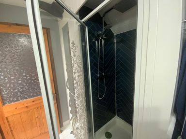 A shower area with dark tiled walls and glass door next to a wooden door in a bathroom at Foxglove Barn in South Brent