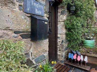 A stone wall with a black mailbox and a door with a knocker next to a shoe rack holding boots at Foxglove Barn South Brent