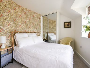 A bedroom with a bed white bedding floral wallpaper bedside table with lamp mirrored wardrobe and a chair by the window at Conybeare Cottage in Lyme Regis