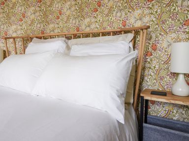 A bed with white pillows and a wooden headboard next to a wooden side table with a lamp in a room with floral wallpaper at Conybeare Cottage in Lyme Regis