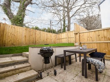 A small patio with a charcoal grill a table and benches next to a wooden fence and steps leading to a grassy area at Conybeare Cottage in Lyme Regis