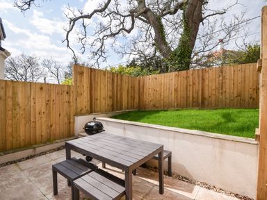 A patio area with a table and benches on a tiled floor next to a grassy raised garden and wooden fence at Conybeare Cottage in Lyme Regis