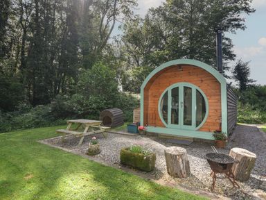 A wooden pod with circular window and green trim in a grassy area with picnic table and fire pit at Pendragon Pod in Axminster