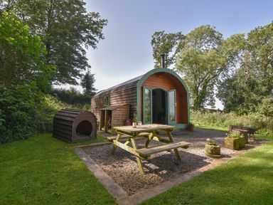 A wooden pod with open doors a picnic table and a dog kennel outside surrounded by trees at Pendragon Pod in Axminster