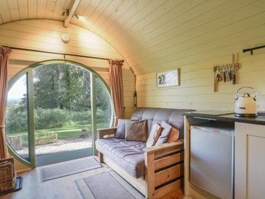 An interior view of a room with a wooden sofa with cushions a small refrigerator a kettle on a countertop knives on a magnetic strip and a round glass door showing an outdoor garden at Pendragon Pod in Axminster