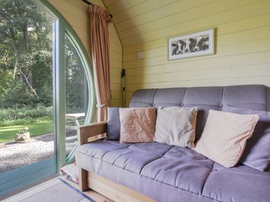 A room with a gray sofa and pillows next to a round glass door opening to a garden at Pendragon Pod in Axminster