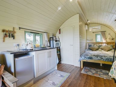 A compact kitchen and bedroom inside a wooden cabin with white cabinets and a bed at Pendragon Pod in Axminster