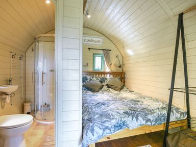 A bedroom with a wooden bed and patterned bedding next to a small bathroom with a shower at Pendragon Pod in Axminster