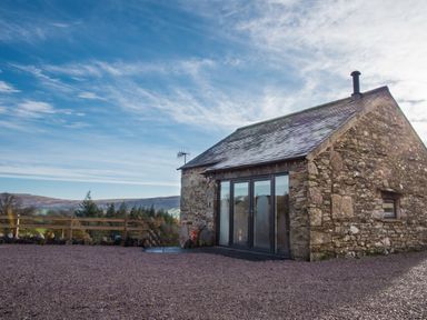 A stone house with glass doors and a chimney on a gravel yard with hills and trees in the background at The Hidden Place in Watermillock near Pooley Bridge