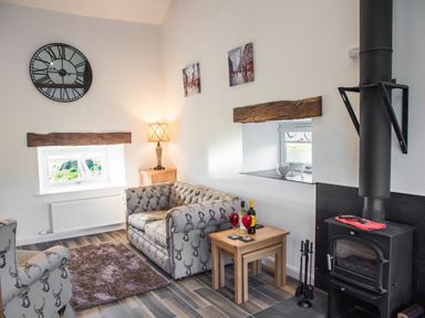 A living room with two armchairs a wood burning stove a side table with wine glasses and bottles a lamp a wall clock and two small windows at The Hidden Place in Watermillock near Pooley Bridge