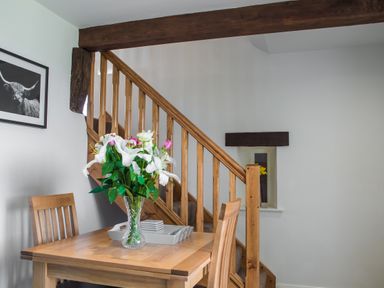 A wooden dining table with a vase of flowers and two chairs near a wooden staircase with framed animal photo on wall at The Hidden Place in Watermillock near Pooley Bridge