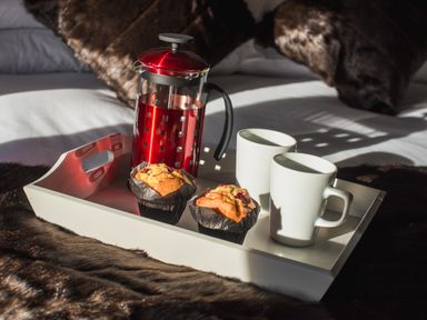 A tray with a red French press, two muffins, and two white mugs on a bed with brown faux fur throws at The Hidden Place in Watermillock near Pooley Bridge
