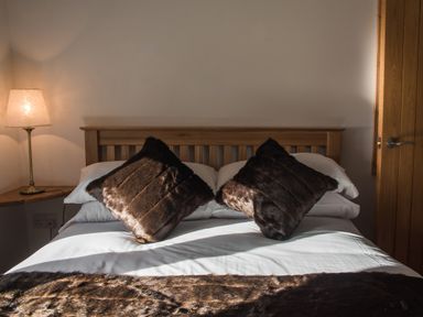 A bed with two dark fur pillows and a fur throw with a wooden headboard and a lamp on a bedside table at The Hidden Place in Watermillock near Pooley Bridge
