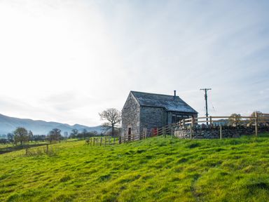 A stone house with a slate roof on a grassy hill with wooden fencing and trees in the background at The Hidden Place in Watermillock near Pooley Bridge