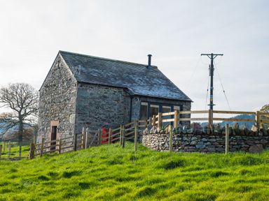 A stone building with a sloped roof surrounded by wooden and stone fences on a grassy hill at The Hidden Place in Watermillock near Pooley Bridge