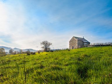 A stone house on a grassy hill with trees and mountains in the background at The Hidden Place in Watermillock near Pooley Bridge