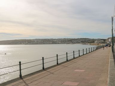 A paved promenade with metal railings along a waterfront and houses on a hill in the distance at Bodillys Cottage in Newlyn