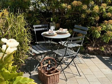 An outdoor patio with a round table set for two with plates and glasses surrounded by four metal chairs and a wicker basket with bottles of wine at Bodillys Cottage in Newlyn