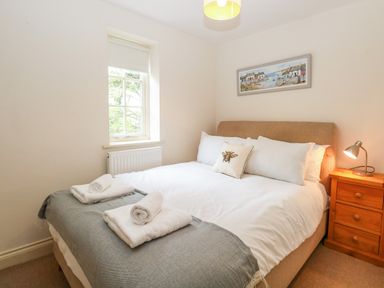 A bedroom with a bed covered with white linens and gray blanket next to a wooden nightstand and window at 30 Burtons Mill in Stalham