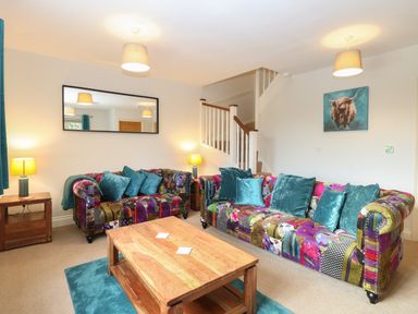 A living room with patchwork sofas, teal cushions, wooden coffee table, side tables with lamps, and a staircase at 30 Burtons Mill in Stalham